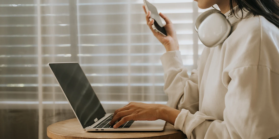 Women wearing headphones and holding her mobile phone in front of her laptop